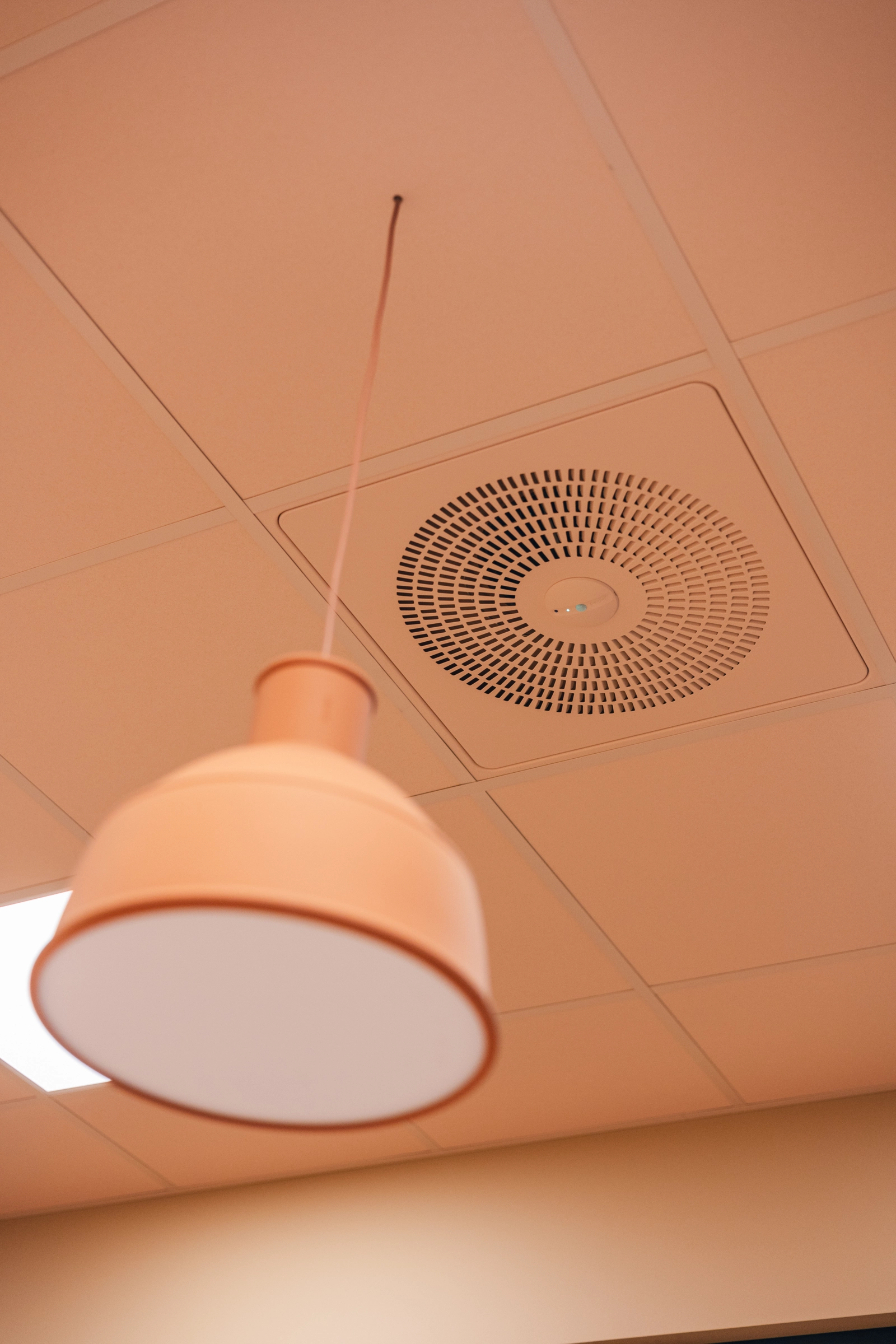 A view of a tiled ceiling in a peach colour with a Swegon WISE Colibri Ceiling perforated air diffuser in the same peach colour